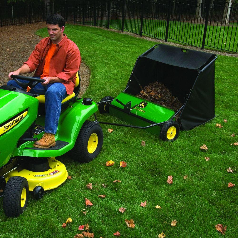 A man in an orange shirt and brown jacket rides a green John Deere lawnmower on grass, towing a John Deere 42 in. 24 cu. ft. Tow-Behind Lawn Sweeper to collect leaves efficiently, with a black fence and trees peacefully in the background.