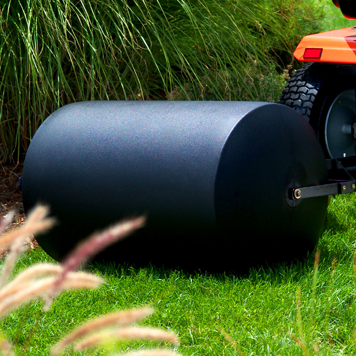 A black Brinly Parts 76 Gallon Tow-Behind Poly Roller | PRT-362BH, 36” wide, is attached to the back of an orange lawn tractor. It rests on green grass with ornamental grasses as a serene backdrop.