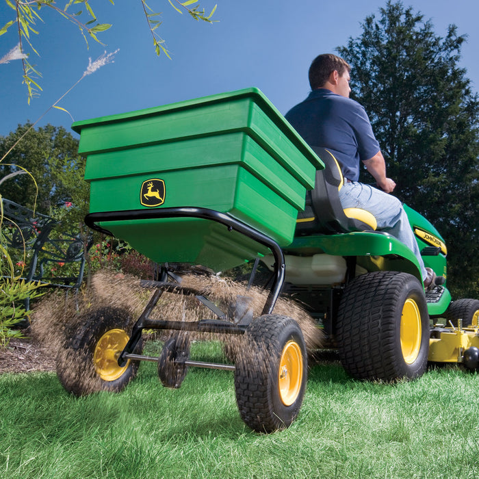 A person skillfully operates a green ride-on lawn mower, pulling a Brinly Parts Tow Broadcast Spreader - 175 lb. Its contents scatter evenly across the lush garden under sunny skies and tall trees, using its pneumatic tires for smooth movement.