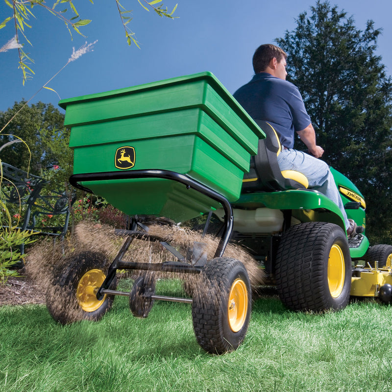 A person skillfully operates a green ride-on lawn mower, pulling a Brinly Parts Tow Broadcast Spreader - 175 lb. Its contents scatter evenly across the lush garden under sunny skies and tall trees, using its pneumatic tires for smooth movement.