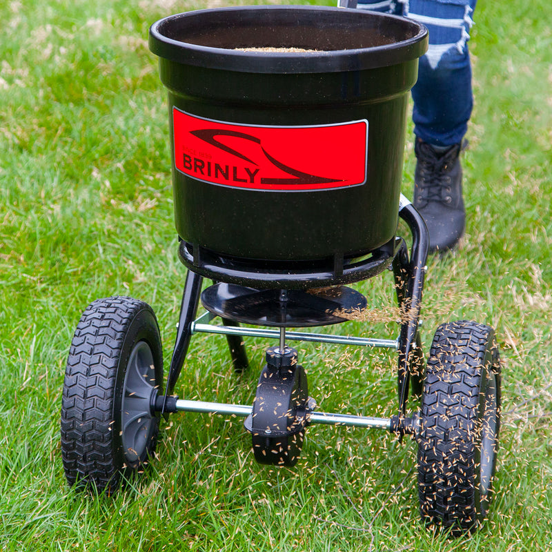 An individual wearing jeans and boots pushes a BRINLY 50 lb. Push Spreader | P20-500BH, evenly distributing seeds across the lawn. The spreader is black with a prominent red label, ensuring complete coverage of every corner.