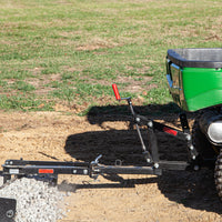 A Brinly Parts green utility vehicle with a gravel spreader attachment efficiently distributes small gravel over a dirt surface. Using a reliable hitch from the Universal ATV/UTV One-Point Lift for Brinly Ground Engaging Products, it is parked on grass, positioned above the gravel pile.