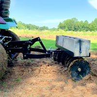 Under a clear blue sky, a tractor with the Brinly Parts Sleeve Hitch Disc Harrow | DD-551BH meticulously prepares soil on a plowed field. Cement blocks enhance harrow efficiency, while trees and grass form a picturesque agricultural backdrop.