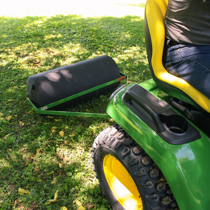 A person uses a John Deere 36 Lawn Roller (PRT-361SJD/LPPRT36JD) with a tethered hitch pin behind a green and yellow ride-on mower. The thick poly drum rolls smoothly over the grass as sunlight filters through the trees, casting dappled shadows on the ground.