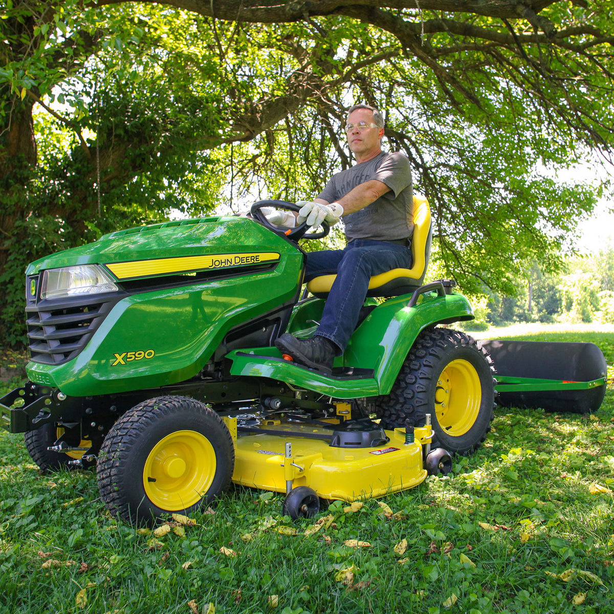 Clad in a gray shirt, jeans, and gloves, a man operates a John Deere 36 Lawn Roller (PRT-361SJD/LPPRT36JD) over the grassy terrain. Sunlight filters through the trees above as he maneuvers with ease, secured by a tethered hitch pin.