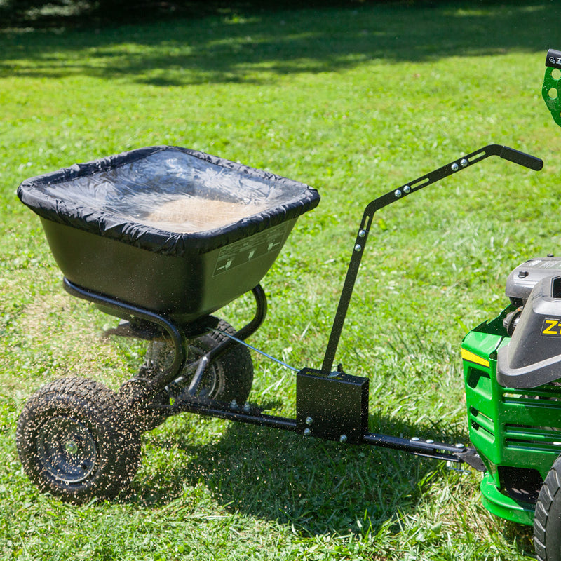 A green riding lawn mower tows a rust-proof Brinly Parts 125 LB. Tow-Behind Deluxe Spreader with Extended Handle & Cover (BS261BH-A) across the grass, efficiently broadcasting light brown fertilizer for optimal lawn care.