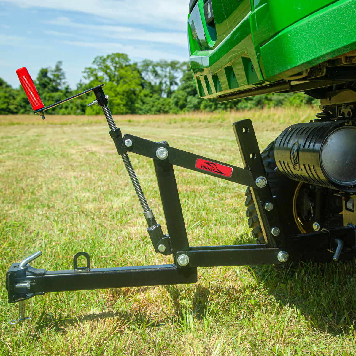 A green tractor equipped with a Brinly Parts Universal ATV/UTV One-Point Lift and a red-handled lever is parked on a grassy field, surrounded by trees against a clear blue sky.