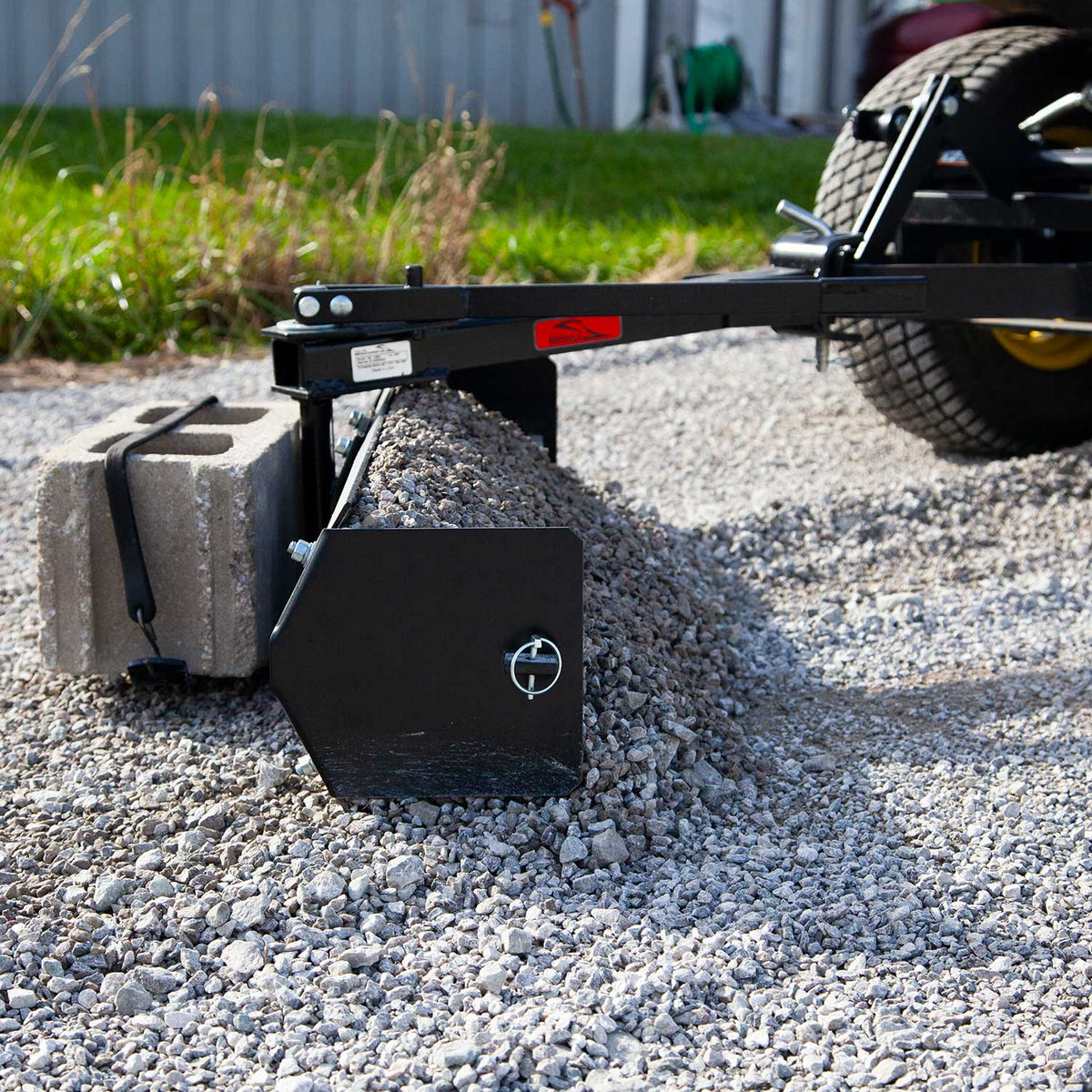 The Brinly Parts 42” Sleeve Hitch Box Scraper (BS-42BH) attached to a lawn tractor effectively spreads gravel on driveways when concrete blocks are added for weight, with grass and a building in the background showcasing this carbon steel blades practicality.