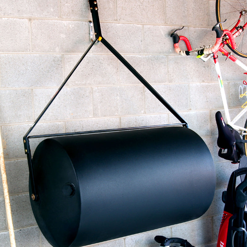 A 76 Gallon Tow-Behind Poly Roller from Brinly Parts is suspended on a brick wall with metal brackets, alongside bicycles and other garage items.