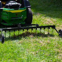 A Brinly Parts 48” Tow-Behind Dethatcher (DT-480BH) is attached to a green lawn mower on the grass. This vital tool features sharp tines for efficient thatch removal, ideal for thorough lawn care. The grass appears to be mid-cleaning and aeration.