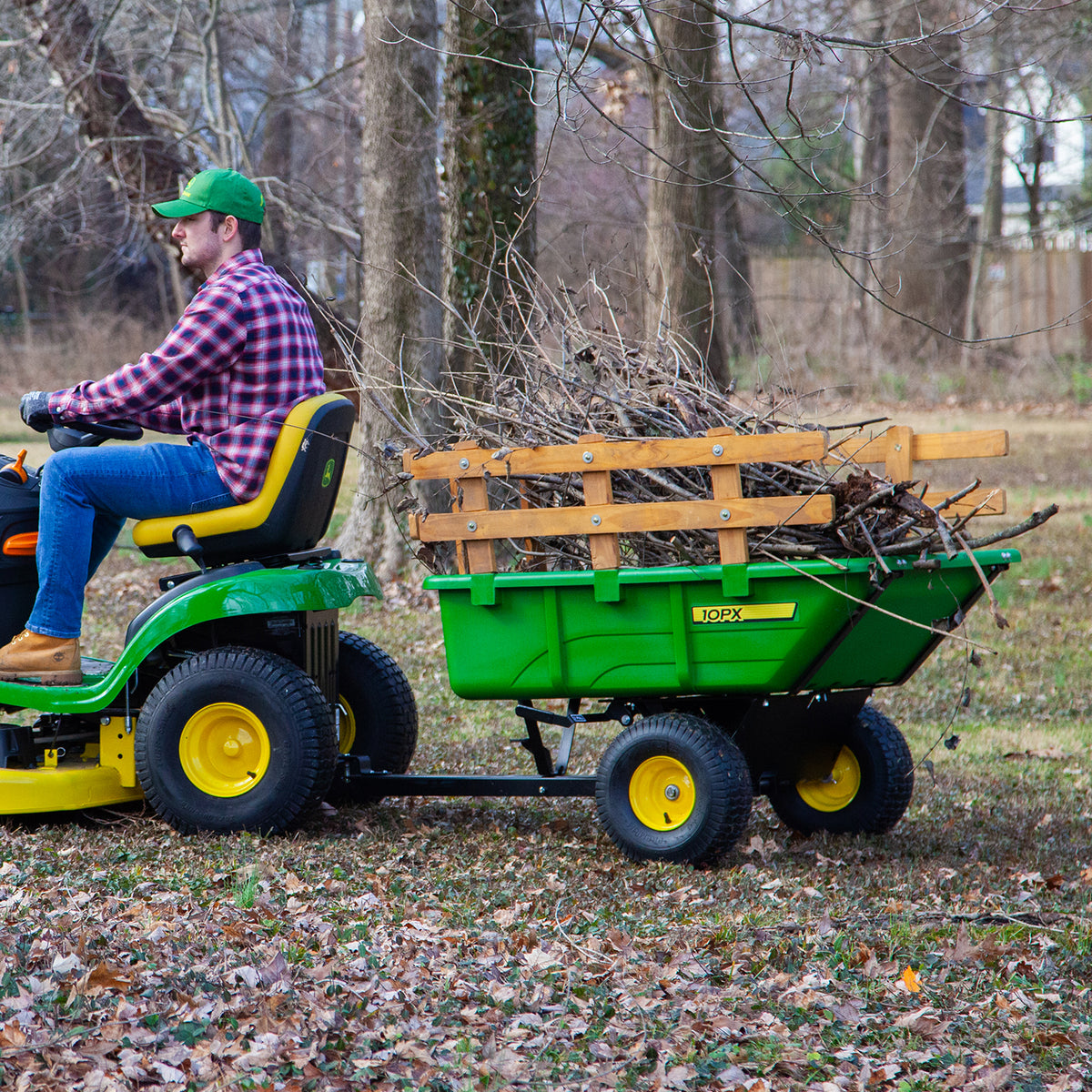 A person in a green cap and plaid shirt drives a green lawn tractor towing a John Deere 180 Degree Full Dump Cart. The cart, with a 650 lb. capacity, is filled with tree branches and twigs. Leaves are scattered across the yard beneath bare trees.