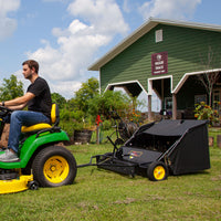 A man uses a Brinly Parts 42″ Lawn Sweeper with Dethatcher (STS-42BHDK-A) on a lawn near the green Veggie Shack building under a partly cloudy sky.