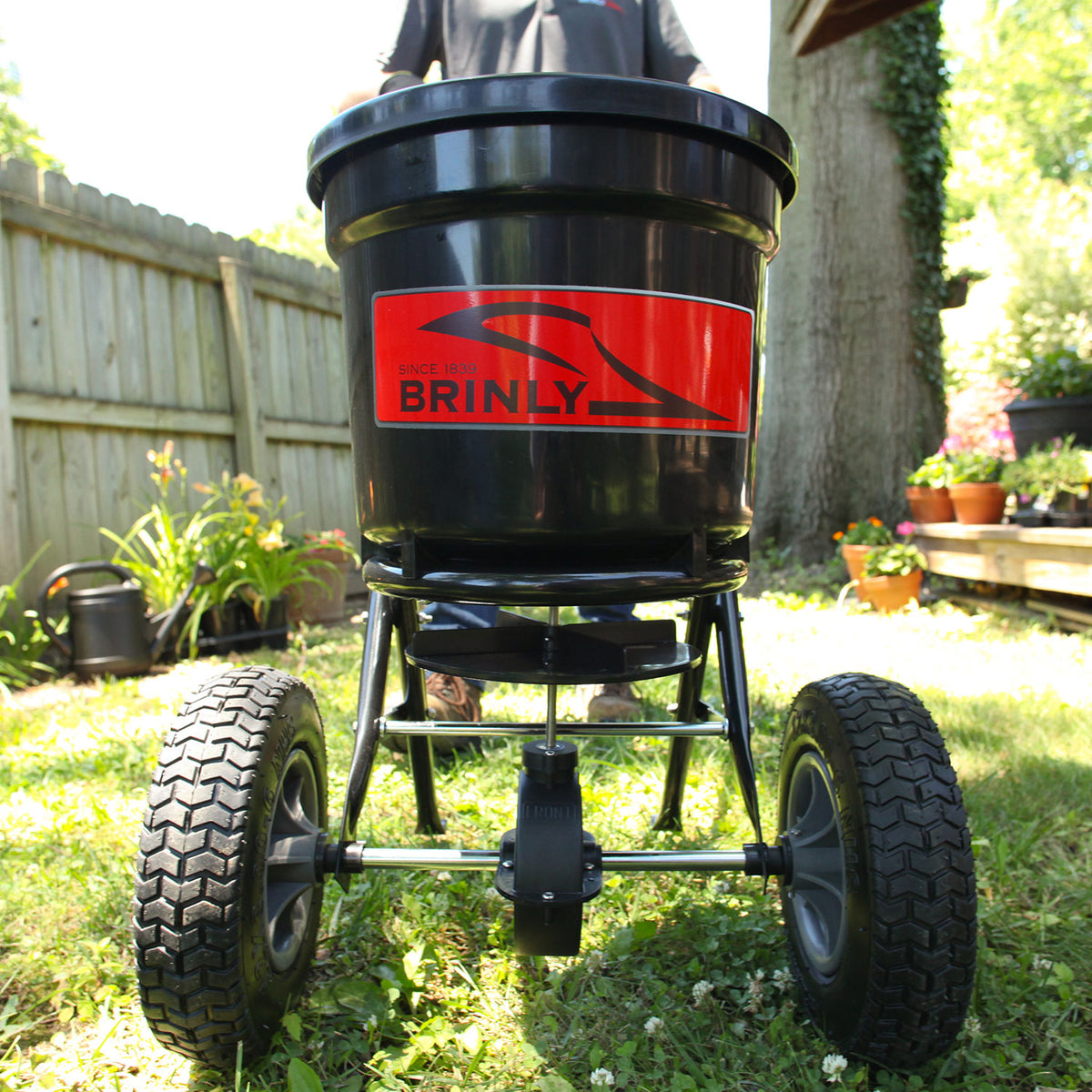 A close-up of a black 50 lb. Push Spreader | P20-500BH by Brinly Parts, featuring a red Brinly logo, rests on grass in a garden. The spreader has two large wheels and is set against a wooden fence with lush plants in the background.