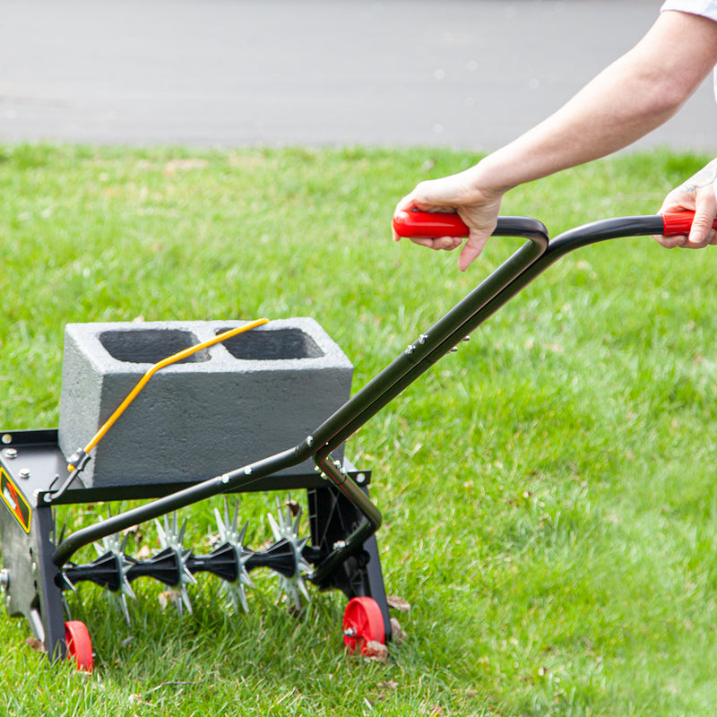 A view of a Brinly Parts 20 Push Spike Aerator with 3D Galvanized Steel Tines on grass highlights its spiked wheels, adjustable handle with red grips, and a concrete block secured on top with a bungee cord for deeper aeration as someone pushes it effortlessly.