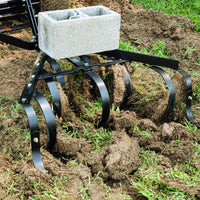 A close-up shows the Brinly Parts Sleeve Hitch Cultivator | CC-560s black metal tines digging into soil, turning grass and dirt for aeration. A concrete block adds weight, enhancing performance similar to a tow-behind cultivator on brown earth mixed with green grass.