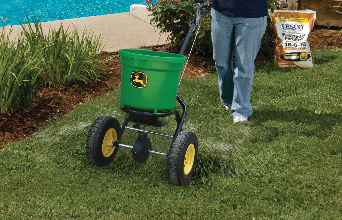 Using a durable poly hopper, a person distributes seed fertilizer over the lawn with a John Deere Broadcast Spreader - 50 lb Round. Nearby, a bag of fall/winter fertilizer waits its turn beside a sparkling pool and a vibrant garden.