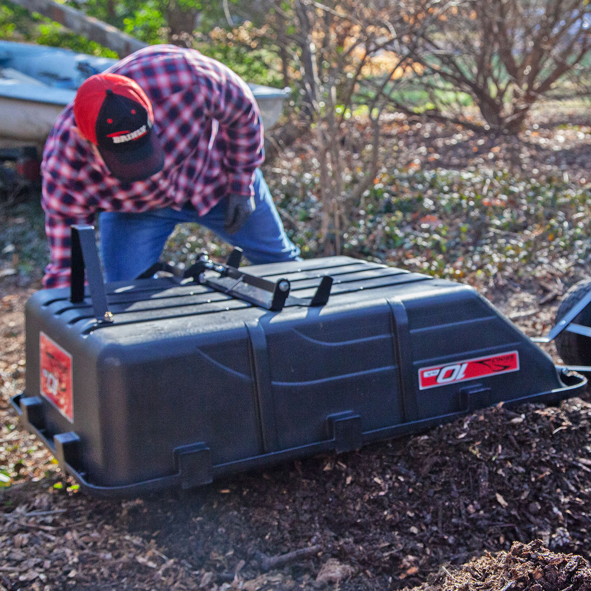 In a red and black plaid shirt and cap, an individual kneels on the ground among trees and mulch, attaching The Roger 180-Degree Full Dump Kit for Brinly-Hardy 10 cu. ft. Poly Carts by Brinly Parts to a riding mower for efficient landscaping.