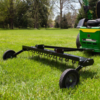 A Brinly Parts 48” Tow-Behind Dethatcher (DT-480BH), with multiple curved tines and two wheels, rests on a green lawn surrounded by trees. The sunny day emphasizes the verdant landscape, exemplifying top-notch lawn care.