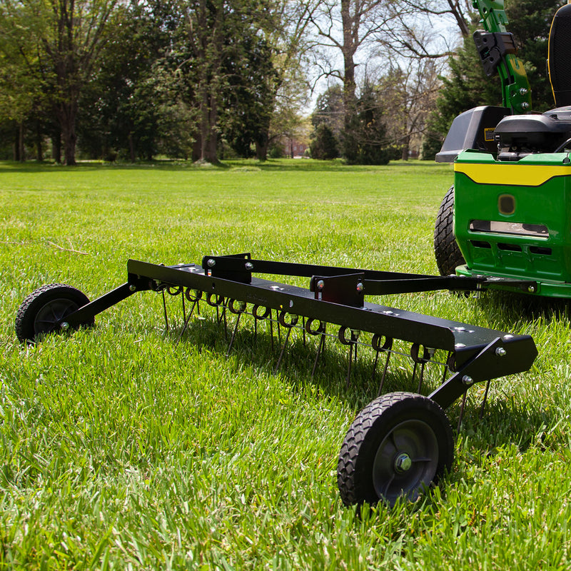 A Brinly Parts 48” Tow-Behind Dethatcher (DT-480BH), with multiple curved tines and two wheels, rests on a green lawn surrounded by trees. The sunny day emphasizes the verdant landscape, exemplifying top-notch lawn care.