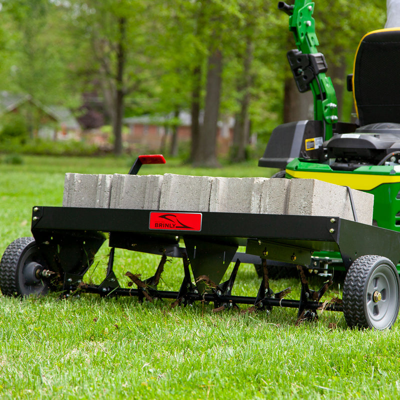 A green lawn tractor is hitched to a black Brinly Parts 48 Tow-Behind Plug Aerator (PA-482BH) filled with concrete blocks. The setup rests on grass surrounded by trees, and the aerator features wheels and steel tines for effective root zone puncturing.