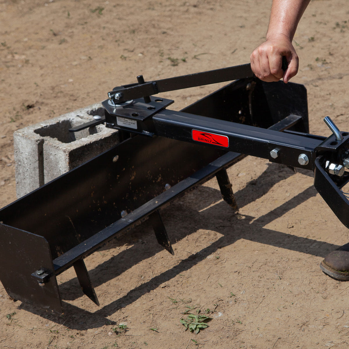 A person adjusts a Brinly Parts 38” Sleeve Hitch Box Scraper (BS-381BH) onto a tractor arm outdoors. The black metal attachment, perfect for grading, is next to a concrete block on the dirt surface and seems indispensable for landscaping or leveling projects.