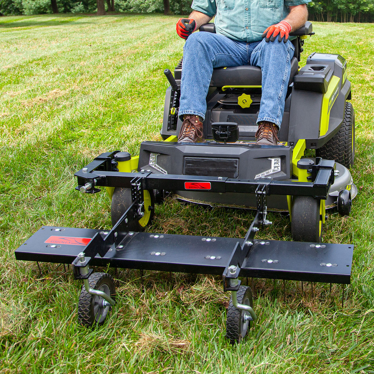 A person operates a 48” ZTR Dethatcher DTZ-481BH by Brinly Parts, expertly mowing a lawn. Wearing jeans, work boots, and gloves, they navigate smoothly. Attached is a black metal accessory with wheels for dethatching. Trees stand in the background.