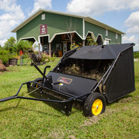 The Brinly Parts 42″ Lawn Sweeper with Dethatcher (STS-42BHDK-A) collects grass clippings on a green lawn outside a green Welcome Center building, featuring yellow wheels and working under a partly cloudy sky.