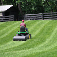 A person in a red cap and plaid shirt drives a green lawn tractor towing a 42 Gallon Tow-Behind Poly Roller (PRT-362SBH) by Brinly Parts across a striped, well-manicured lawn bordered by trees and a black wooden fence on this perfect day, showcasing its 390 lb. capacity.