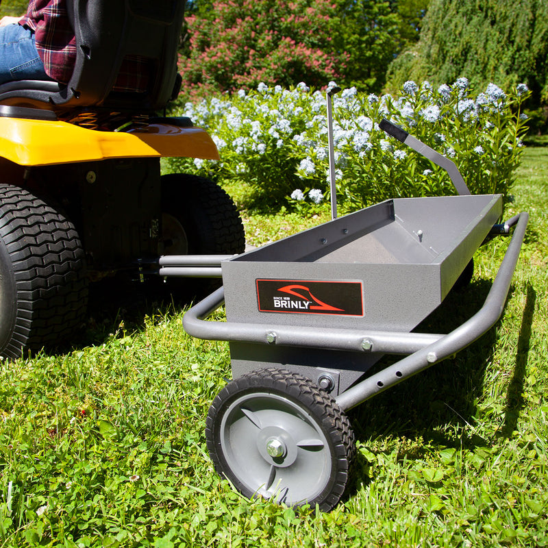 A Brinly Parts 40 Combination Aerator Spreader in Hammered Gunmetal is hitched to a yellow ride-on mower on a vibrant green lawn. Blue flowers and lush greenery in the background suggest a garden or landscaped yard setting.