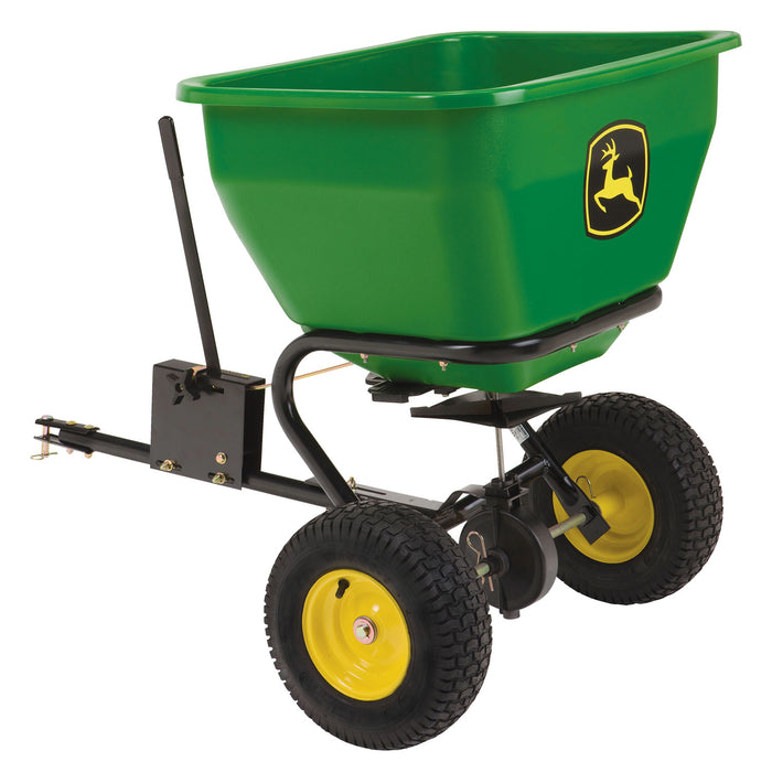 The John Deere Tow Broadcast Spreader - 3.5 cu ft (LPBS36JD) features a green body with a yellow and black emblem, a durable polyethylene hopper, two black wheels with yellow rims, and a metal handle. Its patented directional spread pattern ensures precise distribution when attached to a vehicle.