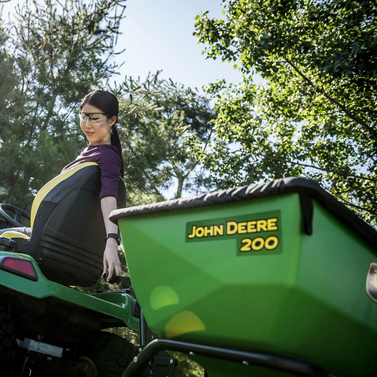 A woman with glasses rides a durable ride-on lawn mower, confidently steering. Wearing a long-sleeve shirt, she gazes back at her green John Deere 200 lb Tow-Behind Broadcast Spreader with ACCUWAY under a clear sky among tall trees.