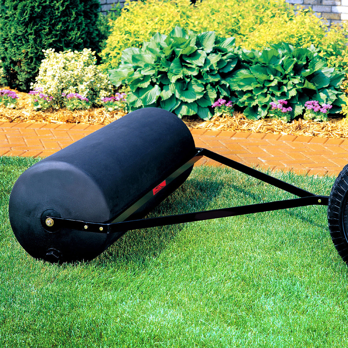 A Brinly Parts 54 Gallon Tow-Behind Poly Roller (PRT-482SBH) with a rust-free poly drum and metal frame handle sits on a manicured lawn, while vibrant gardens and flowering plants flourish in the background bordered by a red brick pathway.