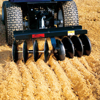 The Brinly Parts Sleeve Hitch Disc Harrow (DD-551BH) with large rotating blades expertly plows the field for soil preparation, creating parallel furrows in the dry, dusty soil while connected to a vehicle via a sleeve hitch.