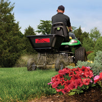 A person rides a green lawn tractor towing a Brinly Parts 125 Lb. Tow-Behind Broadcast Spreader (BS26BH), scattering grass seed on a lush lawn. Pink flowers and shrubs embellish the foreground, with trees under a clear sky in the background.