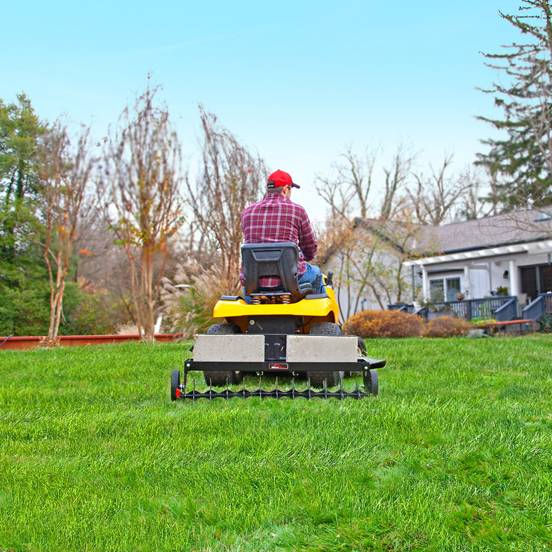 A person in a red cap and plaid shirt rides a yellow lawn mower across the lush green lawn, skillfully maneuvering around their Brinly Parts 40″ Tow-Behind Spike Aerator | SAT2-40BH-G. In the background, trees with sparse foliage frame a house beneath the clear blue sky.