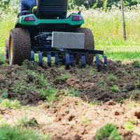 A person uses a green tractor with a Brinly Parts CC-560 Sleeve Hitch Cultivator to till grass and soil. Freshly aerated garden soil is visible in the foreground, with grassy fields in the background.