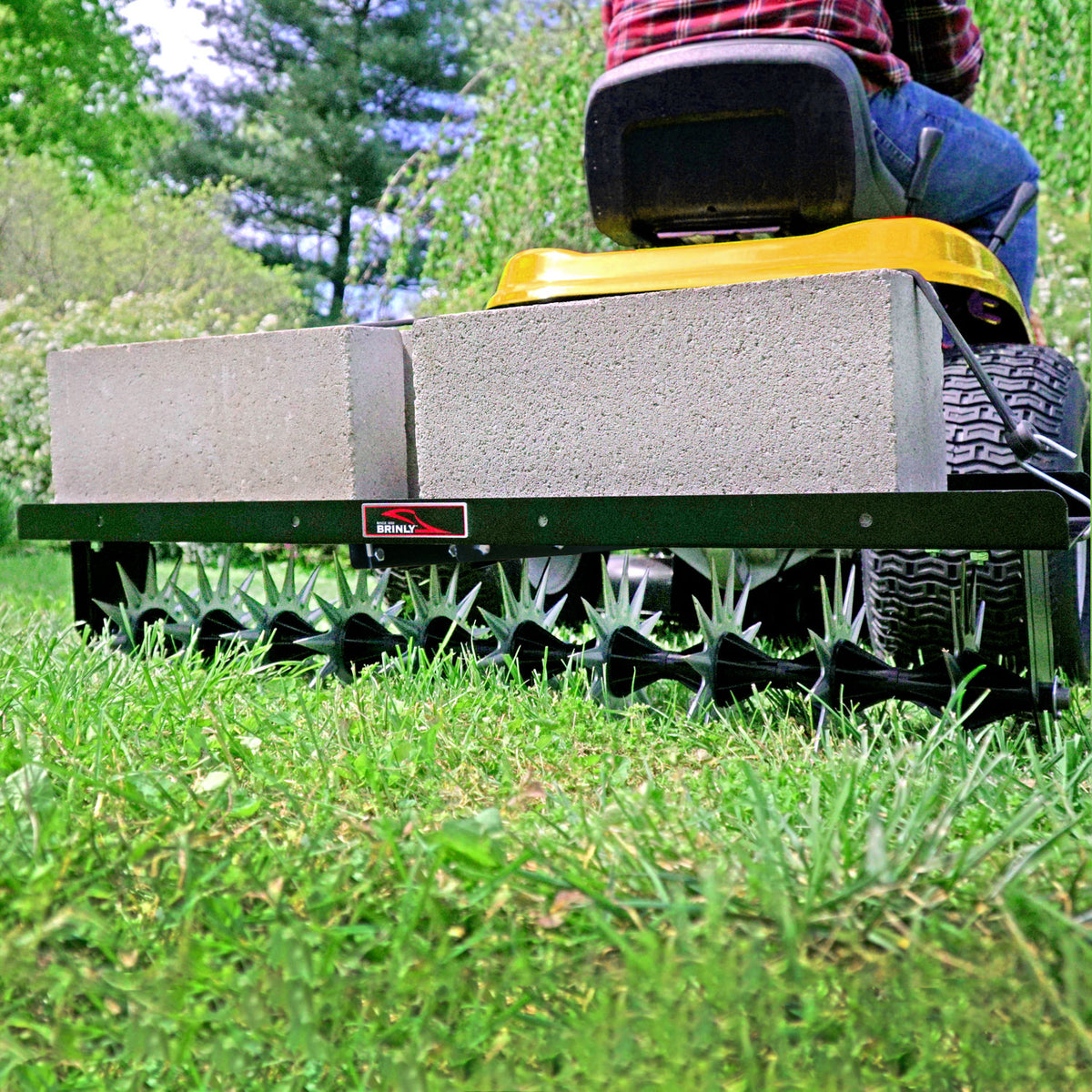 A person uses a yellow lawn tractor to pull the Brinly Parts Spike Aer 40 tow-behind spike aerator, weighted with two concrete blocks, for soil aeration on green grass near trees and bushes.