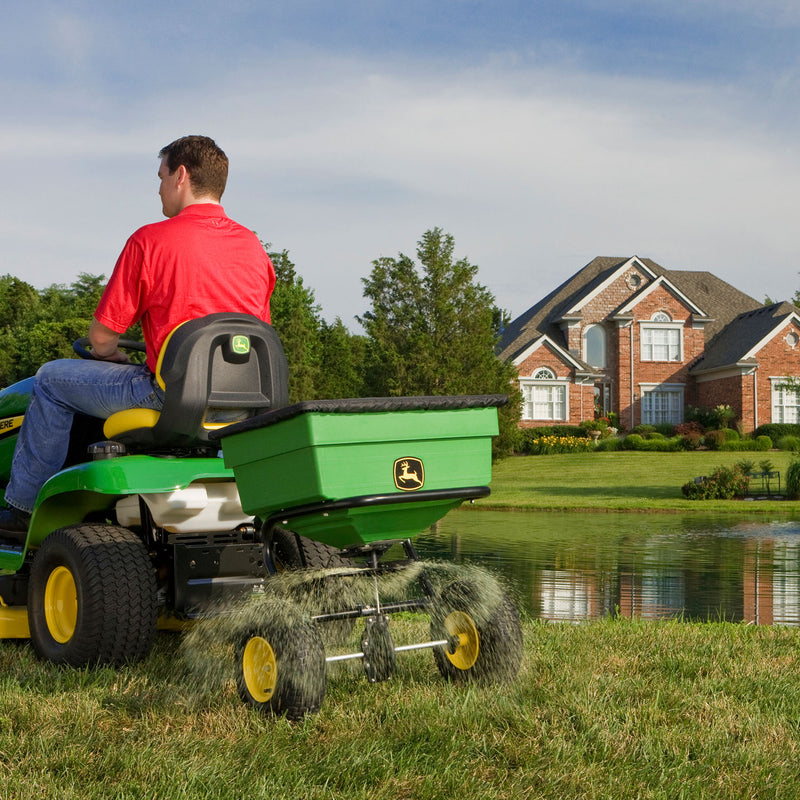 Wearing a red shirt, a person drives John Deeres green Tow Broadcast Spreader - 125 lb near a pond. A brick house with manicured gardens is in the background, while Accu-Way™ Even Spread Pattern Adjustment ensures efficient grass clipping dispersion.