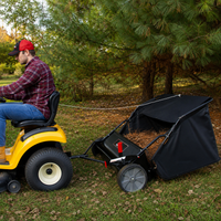 A person drives a yellow ride-on mower towing a Brinly Parts 42″ Lawn Sweeper with DOUBLE-HELIX Brushes (LS2-42BH2-G), which is full of leaves, across a grassy area with pine trees in the background.