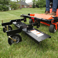 A person in jeans and boots operates an orange zero-turn mower with a Brinly Parts 48” ZTR Dethatcher (DTZ-481BH) attached to aerate a green lawn. Trees and houses in the background highlight diligent lawn care.