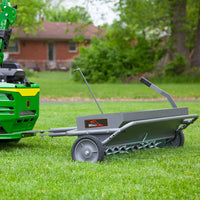 A green lawn tractor tows the Brinly Parts 40 Combination Aerator Spreader AS2-40BH-S in hammered gunmetal across a grassy yard, aerating the lawn with metal tines. A brick house and trees are visible in the background.