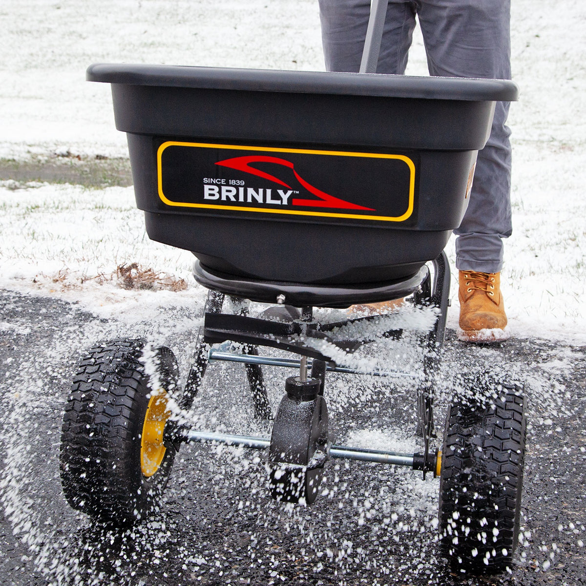 A person in gray pants and brown boots uses the Brinly Parts 70 lbs. Capacity Broadcast Ice Melt Spreader (PS10-70BH) to distribute material on a snowy path, with its calibration dial ensuring an even spray of granules moving across the ground.