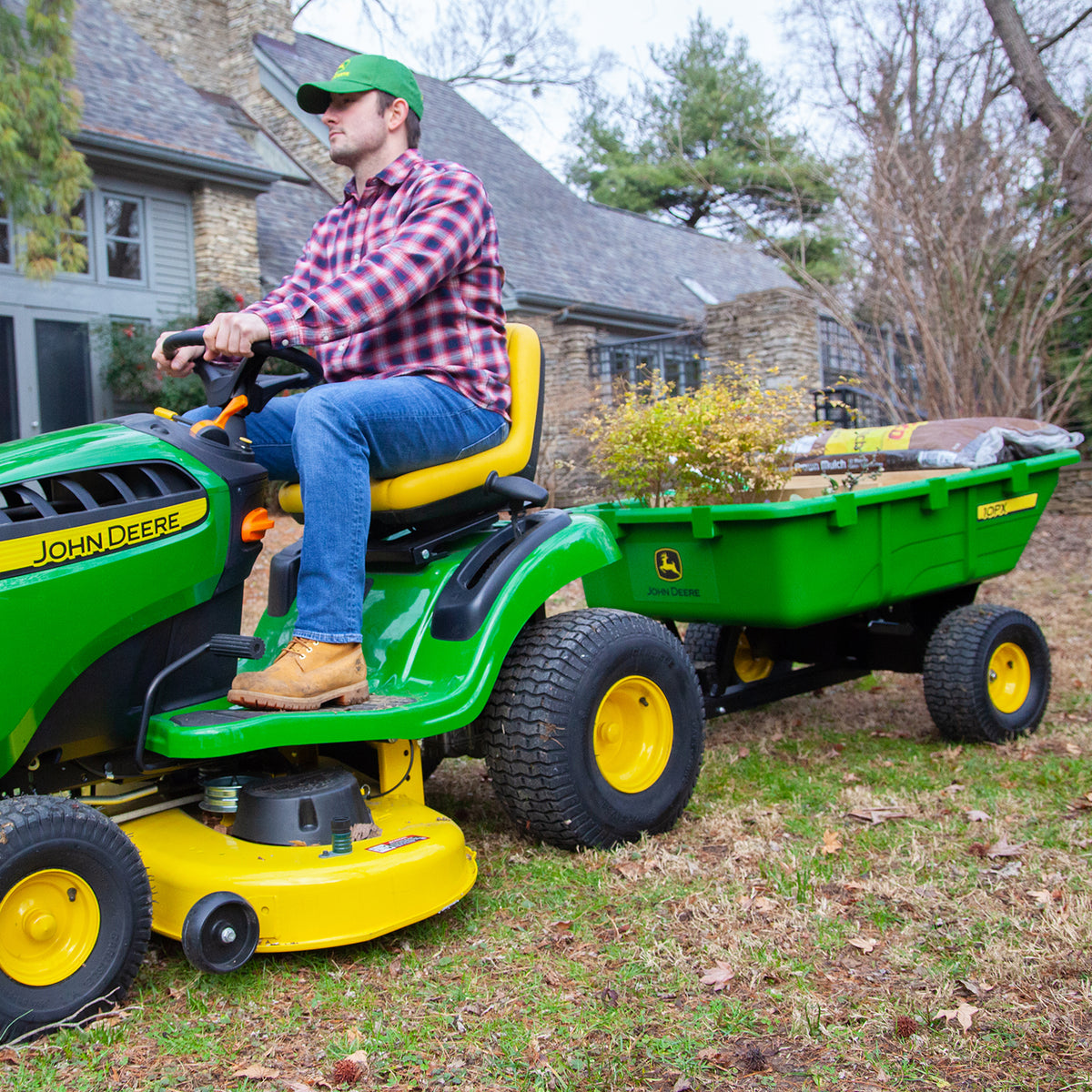 A person in a plaid shirt and green cap drives a green John Deere riding lawn mower towing the 180 Degree Full Dump Cart, which has a 650 lb. capacity, filled with bags of mulch in the yard of a stone house surrounded by trees.