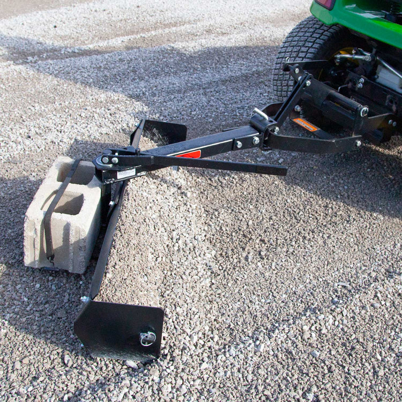 The Brinly Parts 42” Sleeve Hitch Box Scraper (BS-42BH) features a carbon steel blade and adjustable arm. Attached to a green lawn tractor with a cinder block for weight, it efficiently levels gravel driveways.