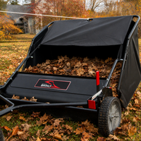 A Brinly Parts 42″ Lawn Sweeper with DOUBLE-HELIX Brushes (LS2-42BH2-G), filled with fallen autumn leaves, sits on the grassy lawn, demonstrating efficient leaf removal amid vibrant trees and a house in the background.
