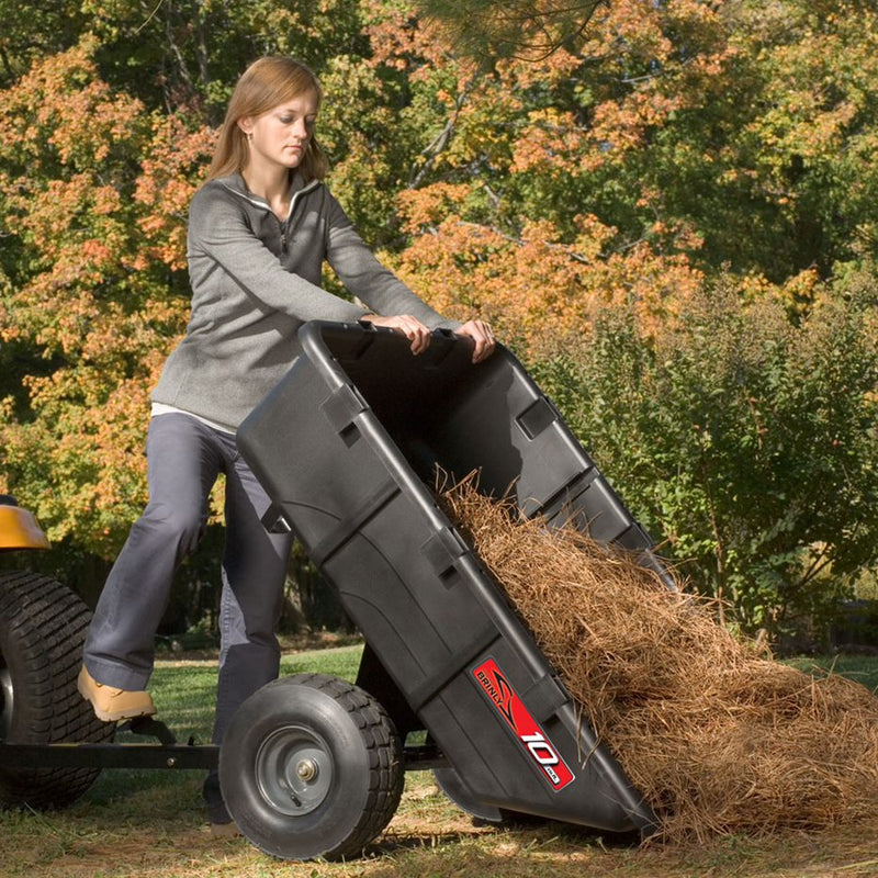 A woman empties dry grass from a Brinly Parts 10 Cu. Ft. Poly Cart (PCT-101BH) in a backyard with trees displaying fall foliage.