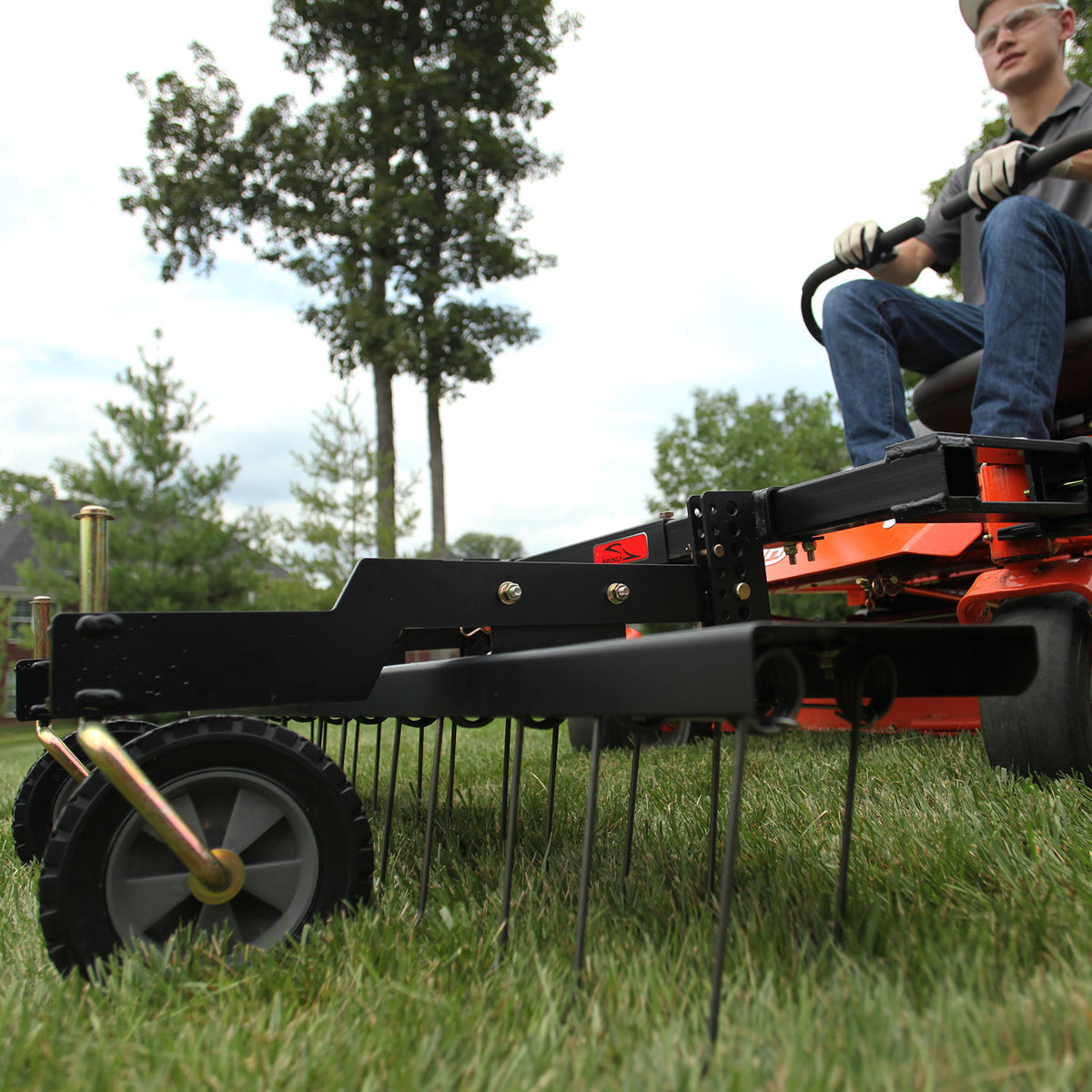 A person skillfully operates a Brinly Parts 48” ZTR Dethatcher, DTZ-481BH, over vibrant green grass. Trees and a house create a peaceful background as they meticulously maintain the lawn, wearing gloves and glasses for protection.