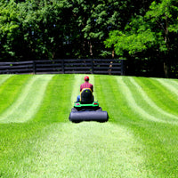 Donning a plaid shirt and red cap, someone guides a riding mower to create neat lawn stripes. Nearby, Brinly Parts 54 Gallon Tow-Behind Poly Roller (PRT-482SBH) enhances the landscape. A dark wooden fence and lush trees enjoy the bright sun.