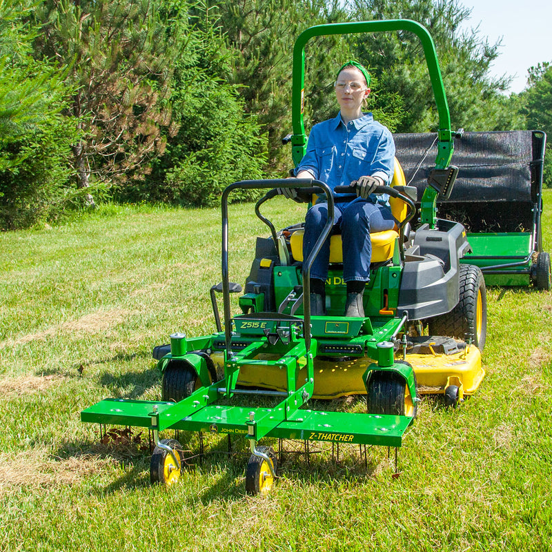 Wearing sunglasses and blue clothes, the person skillfully navigates a John Deere Front-Mount 48 ZTR Dethatcher (CPLP83252). They move effortlessly over the lawn as flexible spring steel tines efficiently work, surrounded by green trees and shrubs on a sunny day.