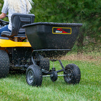 A person on a yellow lawn tractor uses the Brinly Parts BS361BH-A tow-behind spreader with rust-proof polyethylene and a universal hitch to efficiently distribute seed or fertilizer. This black spreader rolls smoothly over green grass on two large wheels amidst trees.